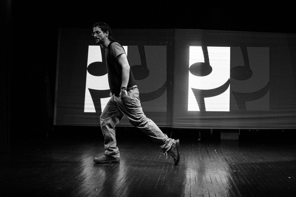 Eduar Campillo en un escenario con cuatro letras musciales de fondo, vistiendo vaqueros desgastados, chaleco de punto, gafas, corte de pelo mallet, barba, pulseras. Da un paso con su pie derecho adelantado y el izquierdo sobre la puntera de sus botas. Lleva chaleco de punto y camiseta de manga corta debajo.